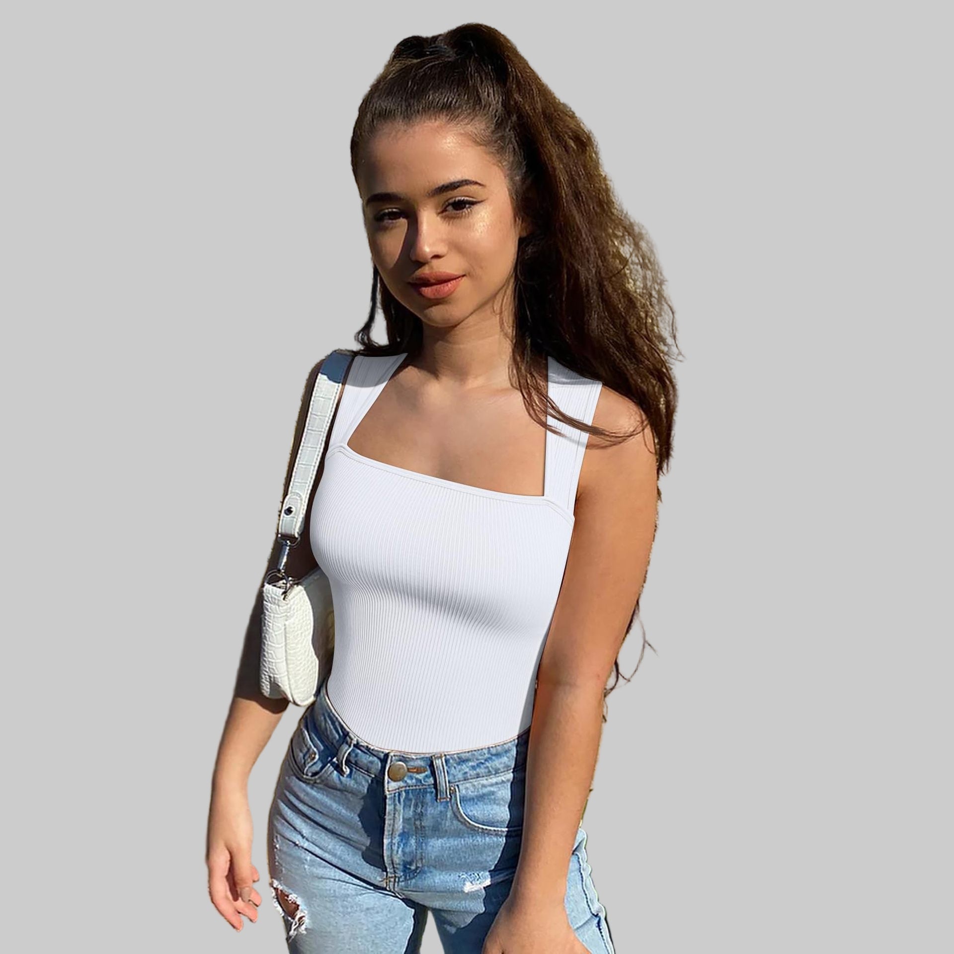 Woman in a white tank top and blue jeans standing against a textured wall.