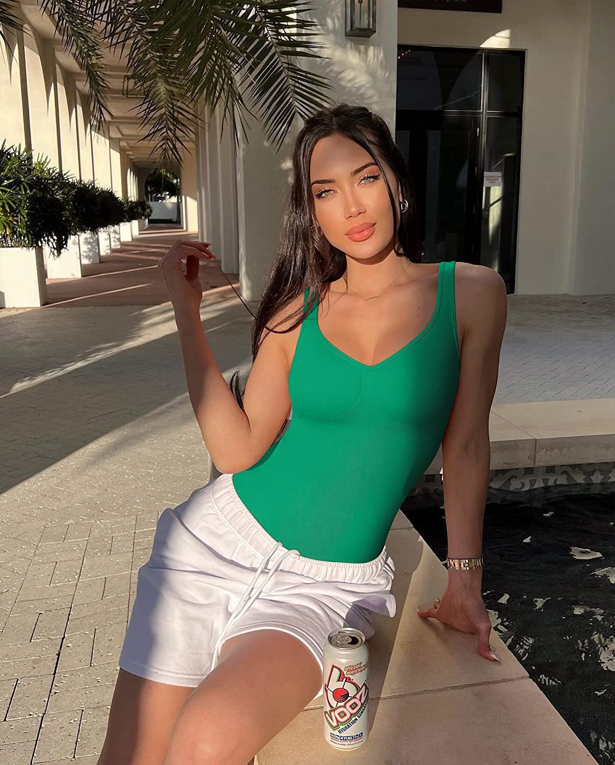 Woman in green swimsuit and white shorts sitting on a bench with a can of Fanta in front of a building.