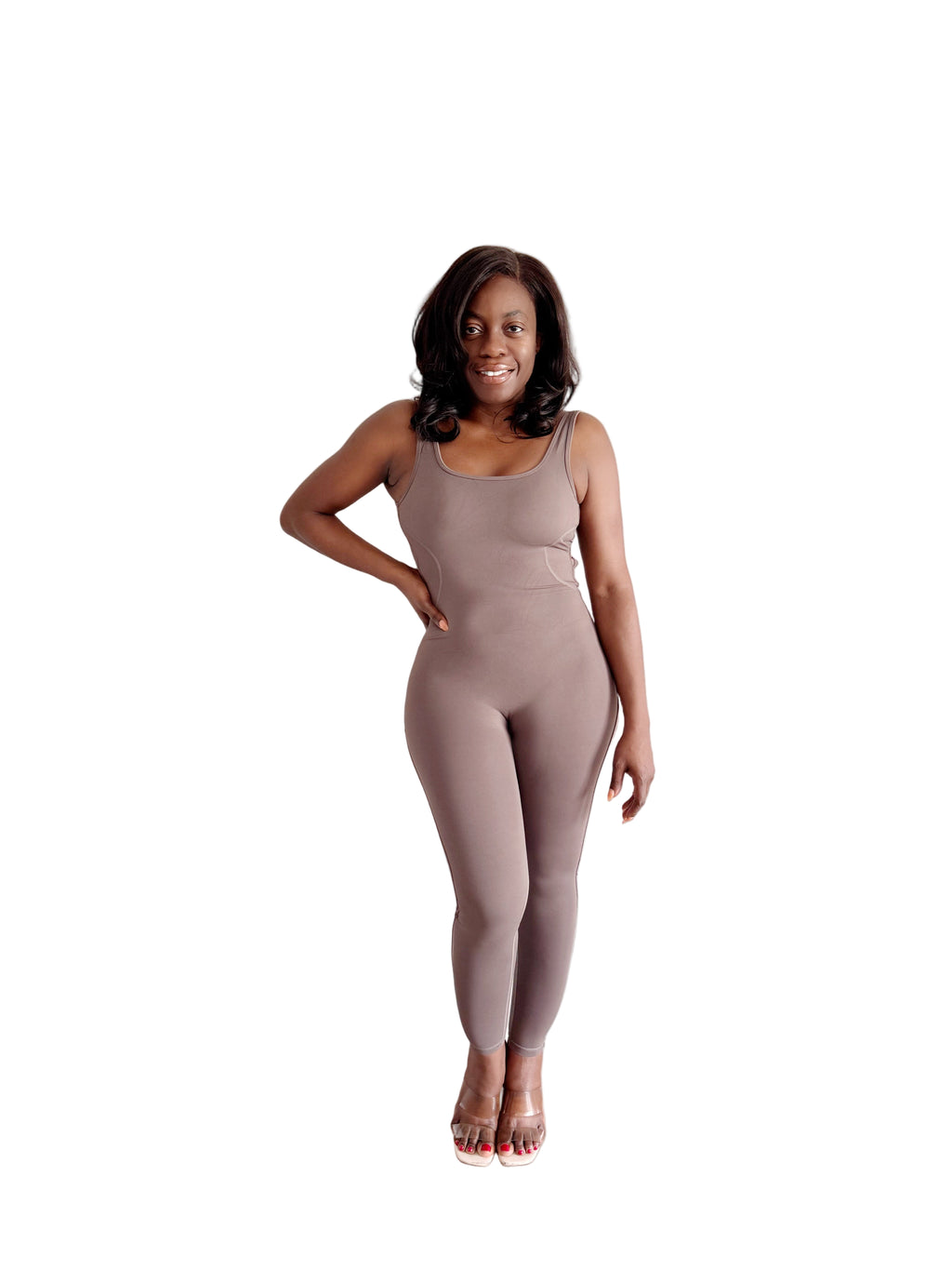 Woman wearing a beige jumpsuit standing in a room with light-colored walls and wooden flooring.
