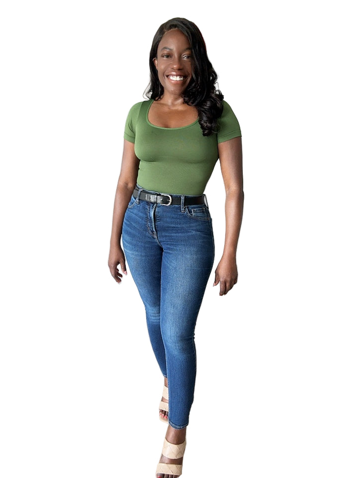 Woman wearing a green top and blue jeans standing in a room with wooden flooring.