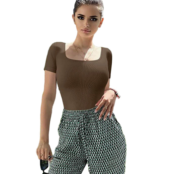 Woman wearing a brown top and patterned pants standing outdoors with greenery in the background