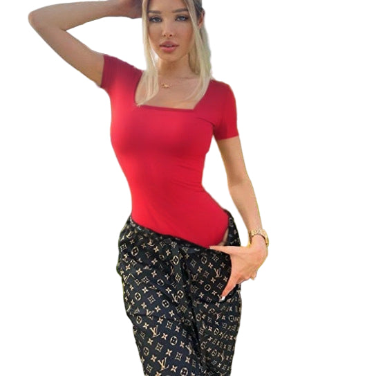 Woman in a red top and patterned pants standing in front of green foliage.
